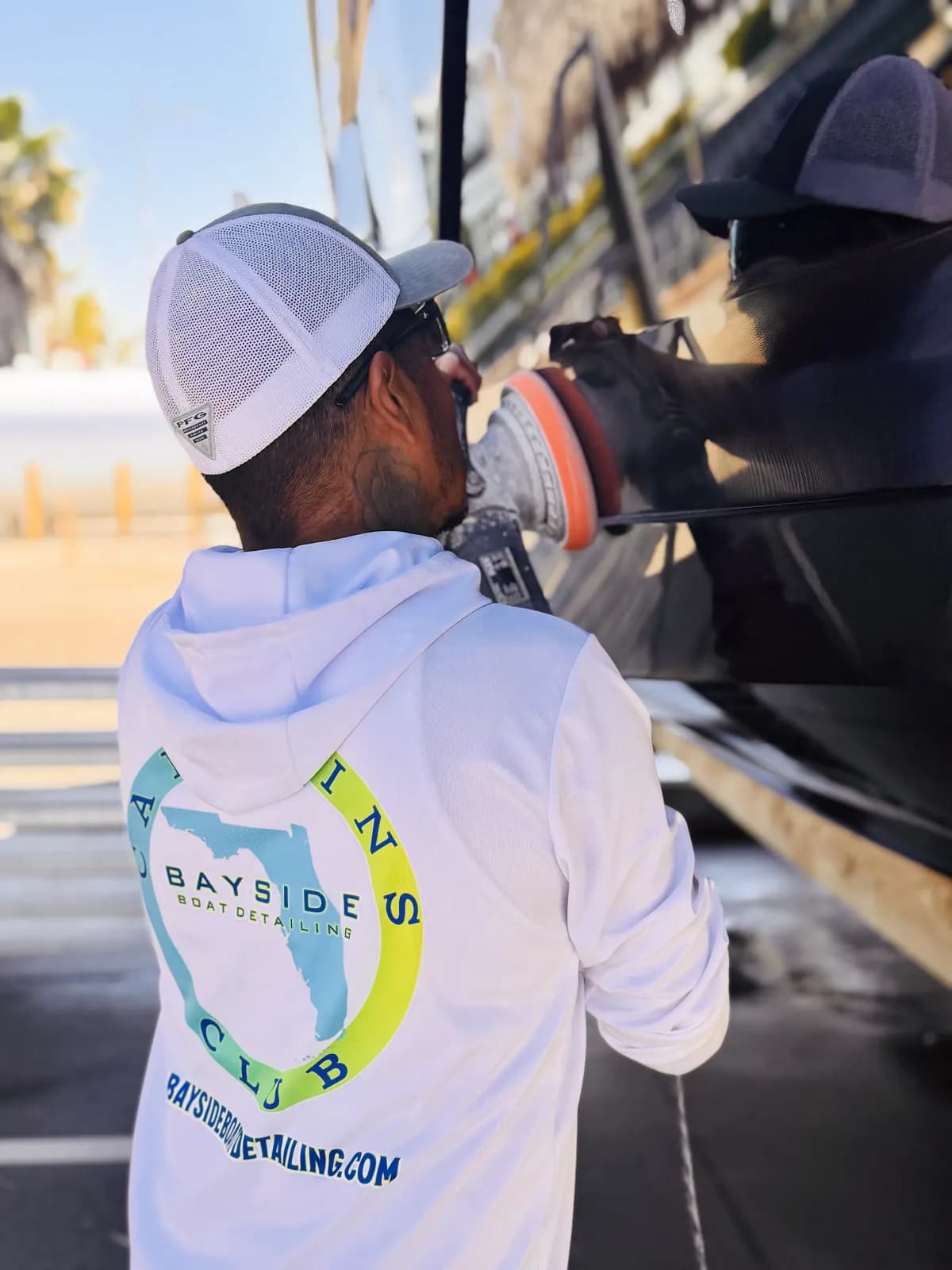 A Bayside technician in a Captain's Club hoodie machine polishing a boat hull in the sun.